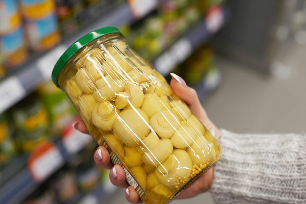a woman is holding a jar of mushrooms in a grocery store, seeking storage tips.